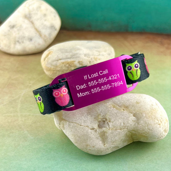 Green and pink owl print kids ID bracelet with pink personalized ID tag displayed on a rock.