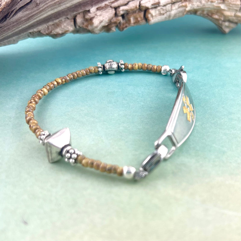 Sand colored and silver beaded medical alert bracelet displayed in front of a piece of wood.