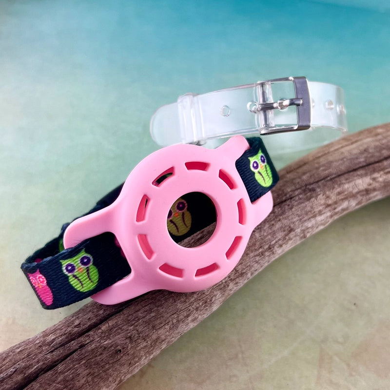 Kids Airtag bracelet in black with printed pink and green owls with pink AirTag holder and clear jelly band displayed on a piece of wood.