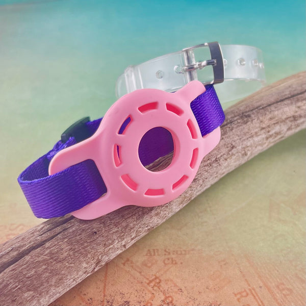 Purple AirTag bracelet holder bracelet with pink AirTag holder and clear silicone bracelet displayed on a piece of wood.