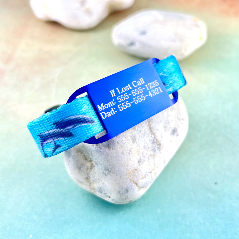 Dolphin print kids ID bracelet and blue ID tag with example of engraving displayed on a rock.