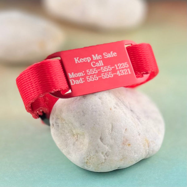 Red nylon kids ID bracelet with custom engraved red ID tag displayed on a rock.