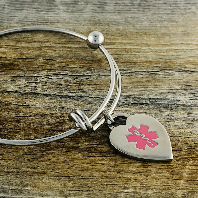 Stainless Steel Bangle and heart medical charm with pink star of life symbol on wood background.