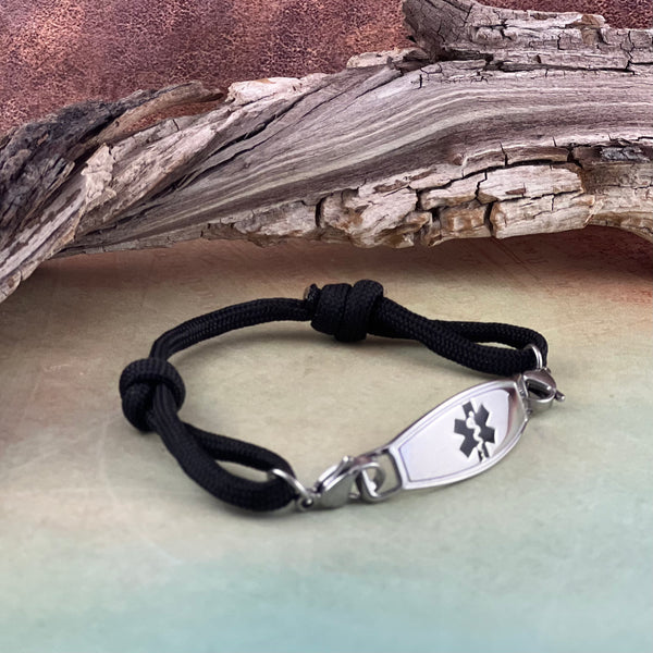 Adjustable black paracord medical alert bracelet with black star of life stainless steel medical ID tag displayed in front of a piece of wood..